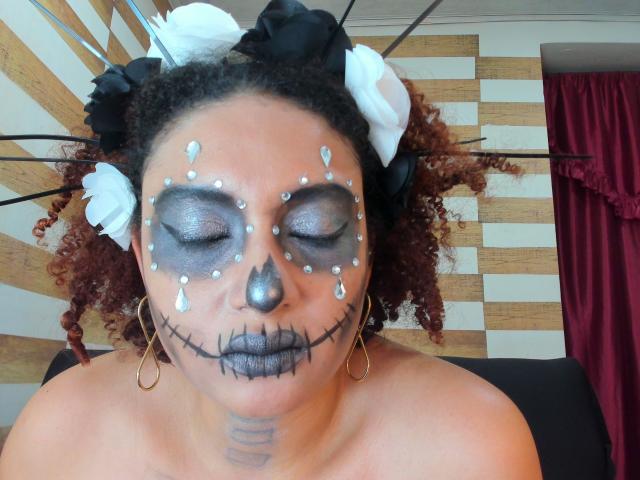 VioletaSwitch heeft donker krullend haar, witte en zwarte bloemen in haar haar, zilveren oogmake-up, zwarte traandruppels, grijze lippenstift, een zwart gestikte mond en gouden oorringen..
