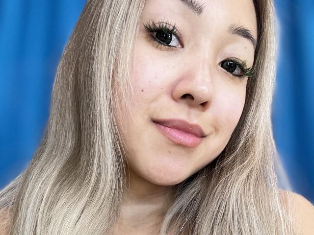 RenyLime has blonde hair, green eyes with long eyelashes, pink lips, and a slight smile. Her face shows a few small moles. She looks cute and confident.