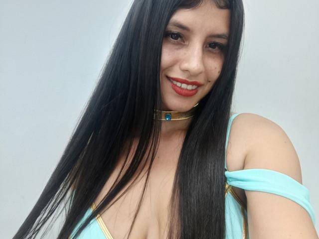 MadissonFontaine with long black hair, smiling, wearing a turquoise dress with gold trim, and a gold choker with a blue gem, showing her tits.
