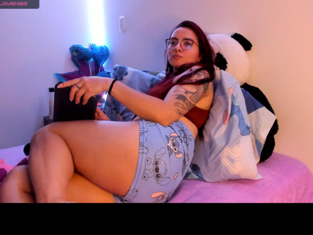 EimmyHoskX with glasses, red hair, tattooed arm, brown top, blue shorts with skull pattern, lying on bed, holding tablet, blue and white pillow, pink bedsheet, soft blue light.