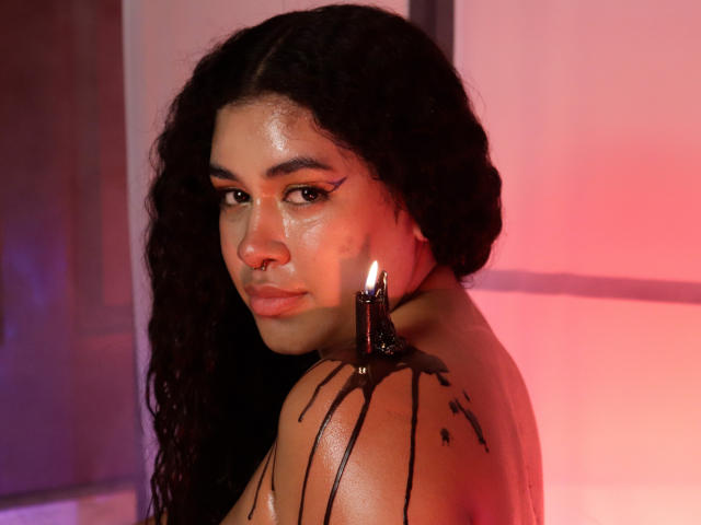 LianaAndAsaff tiene el cabello oscuro y rizado, un piercing en la nariz y un pequeño tatuaje de una estrella en el hombro, con chocolate goteando de una vela en su espalda, mirando a la cámara..