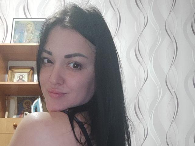 XCrazyBabeX with long black hair, brown eyes, and a slight smile, showing bare shoulders and a black strap, looking over her shoulder. She's in a room with wooden shelves.