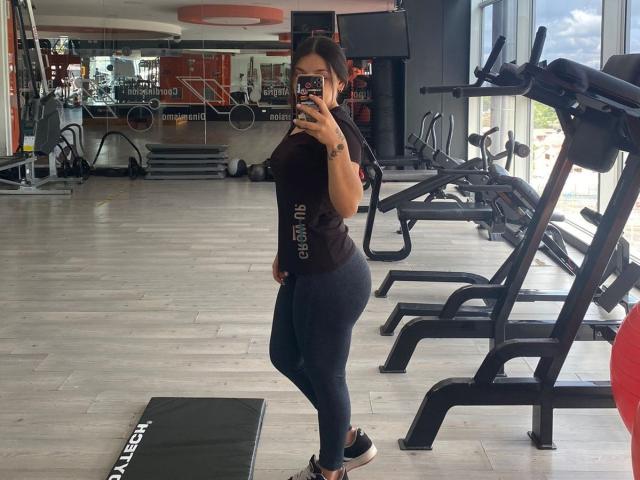 SophiaMarx with curvy ass, wearing black shirt and tight dark blue leggings, taking mirror selfie in gym, holding phone with black case, standing on black mat.