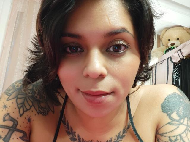 Chili, a sexy Latina woman with shoulder-length black hair, tattooed shoulders, and a necklace, is taking a selfie, smiling softly.