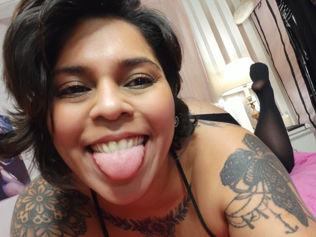 Chili, una mujer tatuada de piel mediana, saca la lengua en una expresión juguetona, vistiendo lencería negra en un dormitorio.