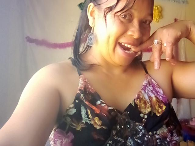 Rahel69, avec des cheveux foncés, souriante, portant une robe fleurie, touchant son menton avec un doigt orné d'une bague, avec des boucles d'oreilles, la lumière du soleil sur son visage, peau brune, seins de taille moyenne, arrière-plan flou..