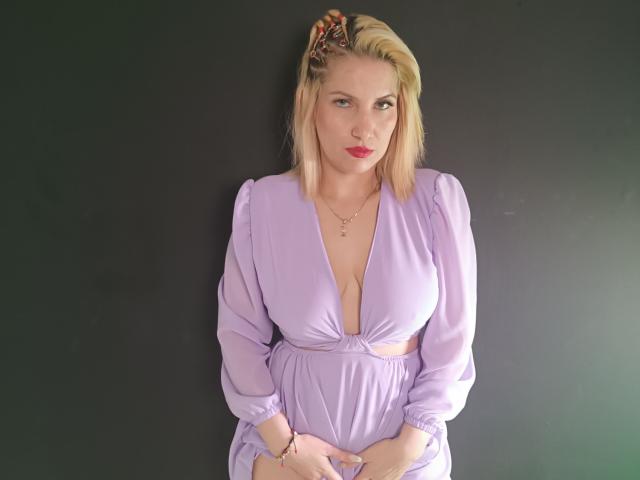 GiuliaAddams with blonde hair and red lipstick, wearing a purple dress with a deep V-neck, showing her big boobs, hands clasped, black background, floral hair clip.