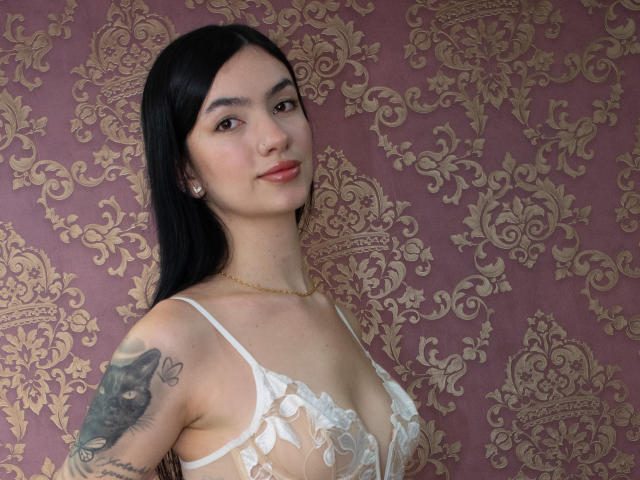 AnnyLondonn with long black hair, wearing a white lace bra, has a tattoo on her right arm, and a slight smile, standing in front of a patterned wall.