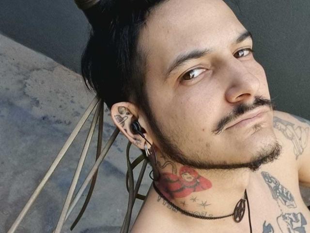 VenusandSata has a top knot, tattoos on the neck and chest, a mustache, ear gauges, and a necklace with a black pendant, looking to the side.