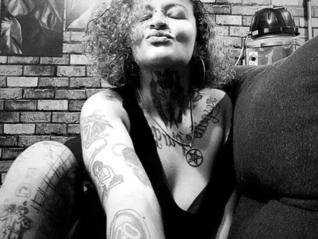 VenusandSata with curly hair, pouty lips, tattoos on neck, chest, and arm, wearing a low-cut top, sitting on a couch, black and white, nipple not visible.