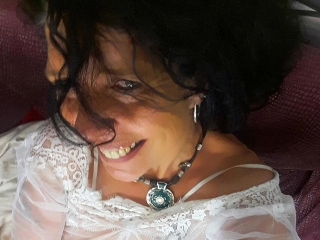 Sexy-Noemy-hot with black curly hair, smiling, wearing a white lace top, and a necklace with a green pendant, showing her teeth and a bit of her nose.