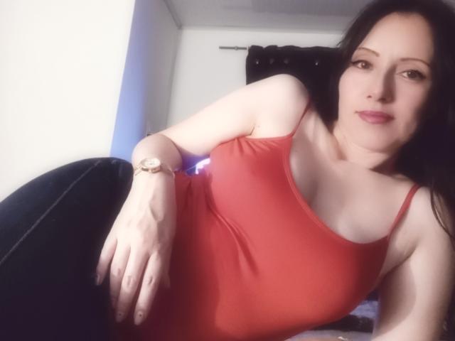 MondBlue with pale skin, black hair, red tank top, and black pants, laying down, looking at camera, wearing a gold watch, slight smile, visible boobs, relaxed.