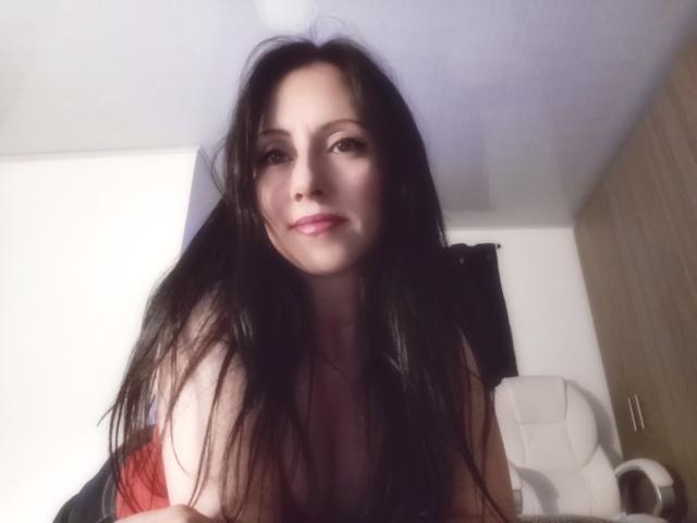 MondBlue with long dark hair, slightly messy, smiling at the camera, wearing a red top, revealing her bare shoulders and upper chest, sitting in a white office chair.