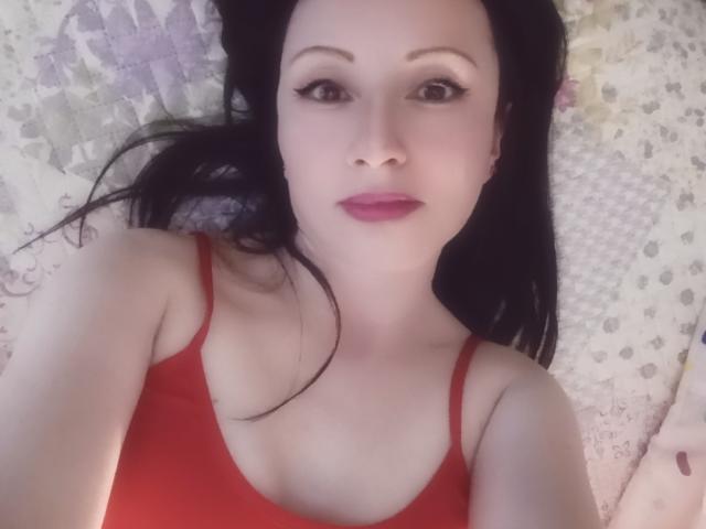 MondBlue has pale skin, black hair, and red lipstick. She's wearing a red tank top, showing her tits, and looking at the camera with a neutral expression.