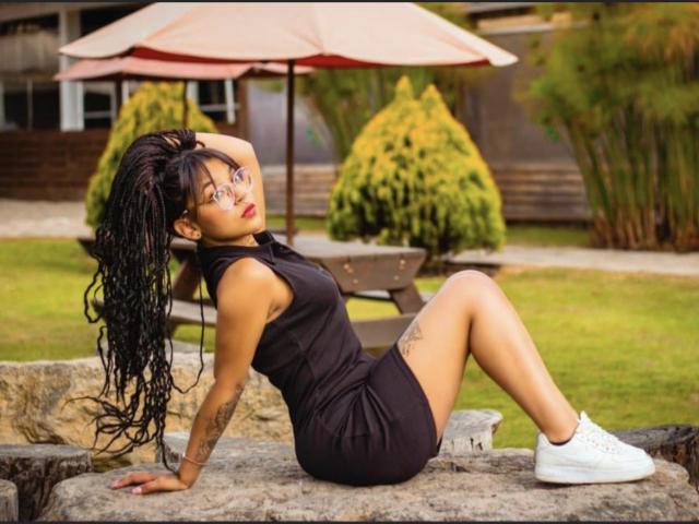DulceTammara69 with long black braids, wearing a black dress, white sneakers, and glasses, sitting on a rock, showing her ass and tattoo, with hands behind her head.
