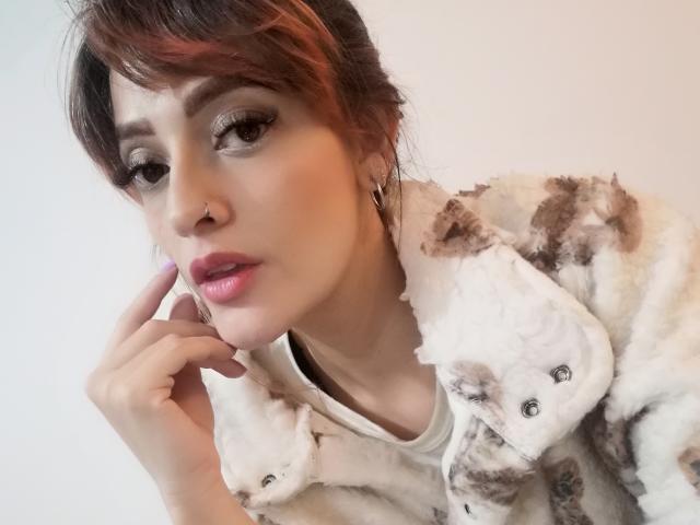 JaneRoa, de piel clara, labios rosados y ojos marrones, vestida con una chaqueta suave de color blanco y marrón con estampado de gato, tocándose la mejilla con la mano derecha..
