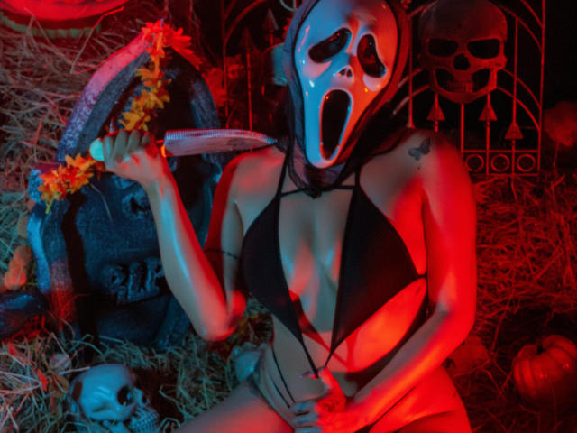 ElizabethMacy in a black bikini, mask with open mouth, holding a knife, red light, hay, skull, and pumpkin, tattoo on right shoulder, hand on her pussy.