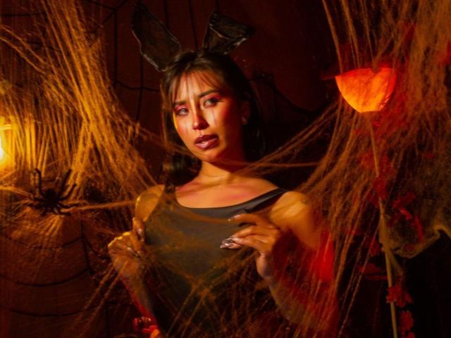 Elizabeth Macy, con cabello oscuro, lleva orejas de conejo negras, una camiseta gris y telarañas, tiene las manos sobre sus pechos, luciendo seductora con iluminación roja y naranja..