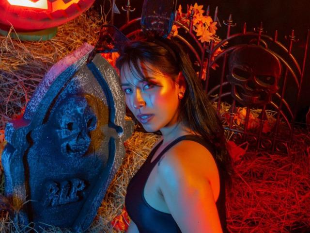 ElizabethMacy with dark hair, wearing a black tank top, looking at the camera with a serious expression, standing next to a spooky stone skull with a bat on her head.