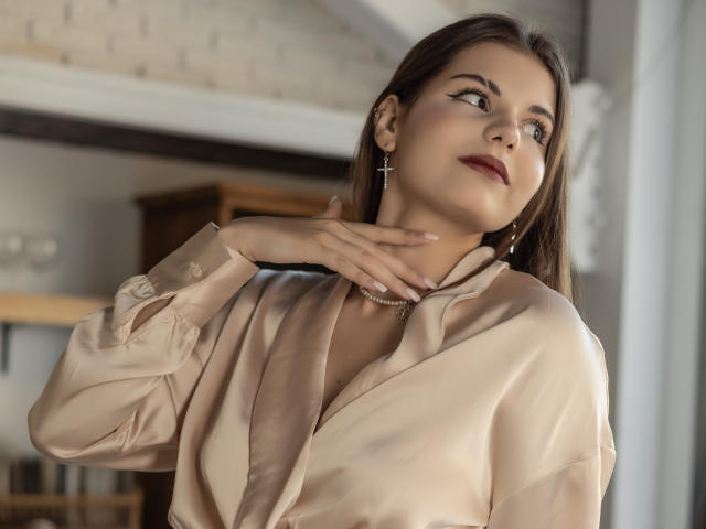 AdeliaLove with dark red lipstick, brown hair, cross earrings, and a beige satin blouse, hand near her neck, looking up, in a soft indoor light.