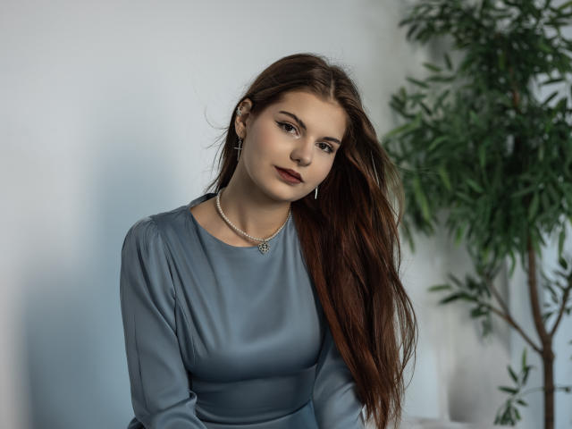 AdeliaLove with long brown hair, wearing a gray dress, small tits, and a necklace, smiling softly, standing next to a green plant, in a well-lit room.
