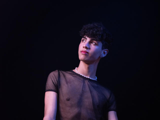 NeoRaptis, avec ses cheveux noirs bouclés, portant un haut en maille noire transparent et un collier de perles blanches, regardant vers le haut avec un léger sourire, fond noir, avec des reflets violets clairs sur le visage..