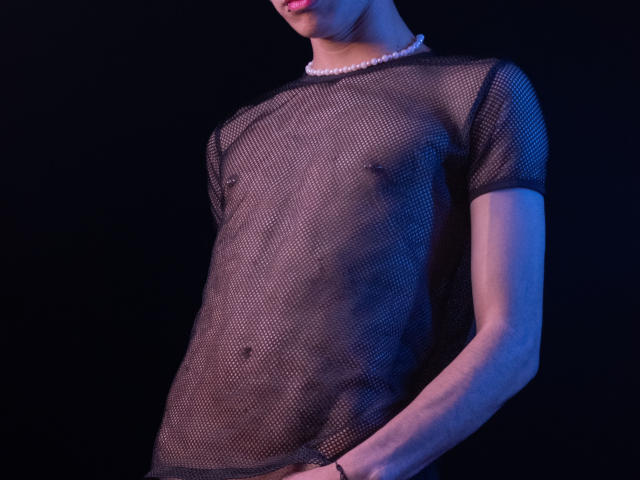 NeoRaptis wearing a black mesh shirt, showing a flat chest and visible nipples, with a pink pearl necklace, against a dark blue and black background.