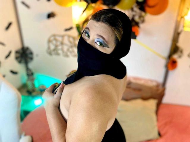 Salma Azaman, con maquillaje de ojos verde, un pañuelo negro que le cubre la boca y la nariz, dejando al descubierto sus hombros desnudos y un vestido negro, está de pie con la espalda hacia la cámara..