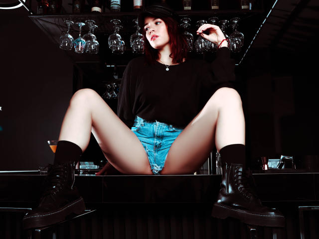 MiaTabasco, legs spread, black boots, blue shorts, black top, black hat, sitting on bar, glass bottles above, hands in hair, looking up, white skin, red lipstick.