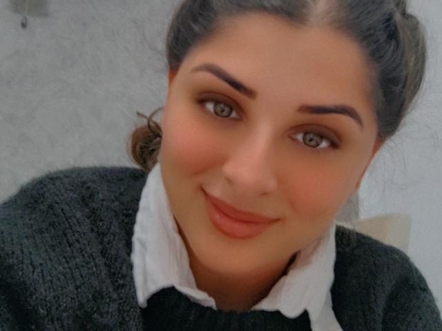 ScarletOra has brown eyes, full eyebrows, and a subtle smile. She's wearing a gray sweater with a white shirt underneath, her dark hair in a messy bun.