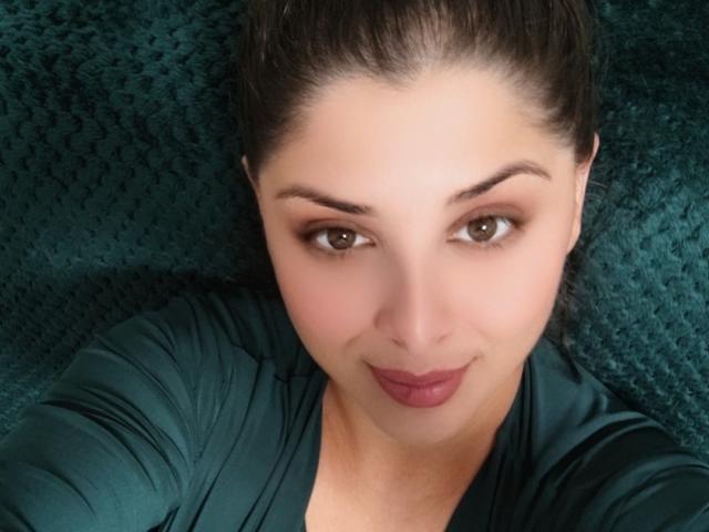 ScarletOra has her dark brown hair pulled back, wearing a teal shirt, with her brown eyes and pink lipstick, lying on a teal, textured fabric, smiling slightly.