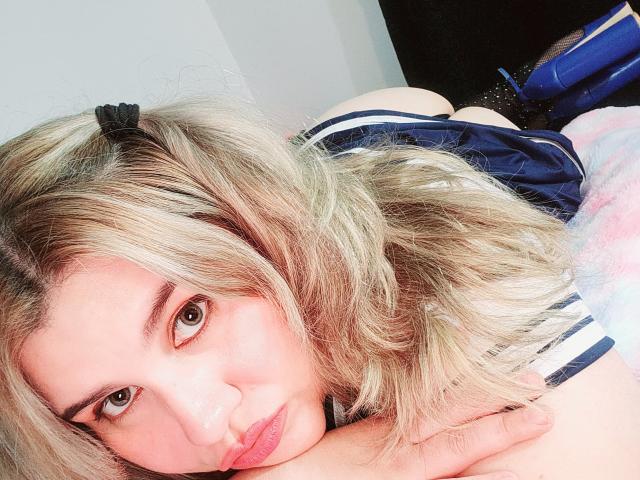 Akkanee has blonde hair, green eyes, and is lying on a bed with her face near her ass, wearing a navy and white striped top. She's pouting slightly.