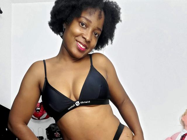 ChelseyeBonny, con piel oscura, cabello negro rizado y lápiz labial rosa, vestida con un sujetador y tanga negros de Calvin Klein, sonriendo a la cámara, mostrando sus pequeños senos..