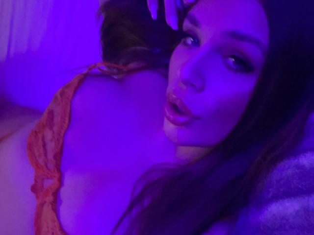 IvyRowa with purple-lit face, wearing orange lace bra, exposing her big boobs, has long brown hair, slightly open mouth, looking at camera, in soft focus.