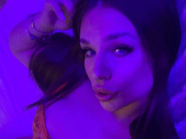 IvyRowa with purple-lit face, dark hair, and makeup, looking at camera, slightly parted lips, right hand raised, wearing red lace top, bracelet on wrist.