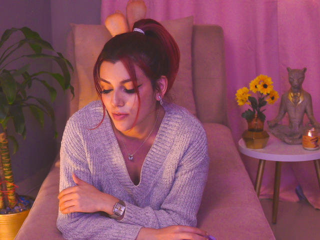 ZoeeMar with red hair in a ponytail, wearing a gray sweater, crossing her arms, sitting on a beige couch, purple and pink lighting, small round table with candles.