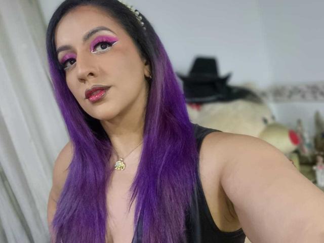 AndreaSharick with purple hair, pink eyeliner, and glossy pink lipstick, wearing a black tank top and gold necklace, smiling at the camera. Boobs visible, cute and sexy.