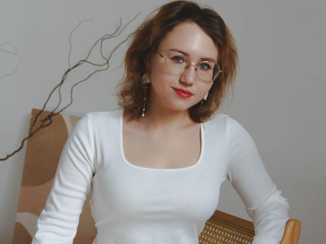 TeresaBirt with curly brown hair, red lipstick, glasses, white long-sleeve shirt, and earrings, sitting on a wicker chair. Boobs slightly visible under shirt.