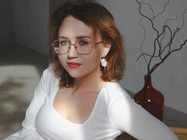 TeresaBirt has light skin, blue eyes, and brown wavy hair. She's wearing glasses, a white long-sleeve top, and diamond earrings, with red lipstick. Boobs are visible.