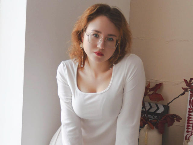 TeresaBirt with wavy red hair, glasses, and a white long-sleeve top, leaning forward, showing her small, perky boobs. Soft light, slightly blushed cheeks, subtle makeup.