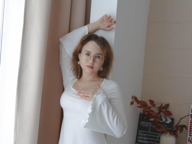 Teresa Birt, usando óculos, batom vermelho e uma camisa branca de manga comprida, com uma mão na parede e a outra no peito, cabelo castanho ondulado, em pé perto da janela, olhando para a câmera..