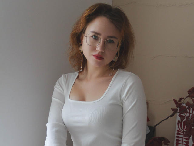 TeresaBirt with wavy red hair, glasses, and white long-sleeve top showing her tits, standing near a wall, red plant in the background. She's looking at the camera.