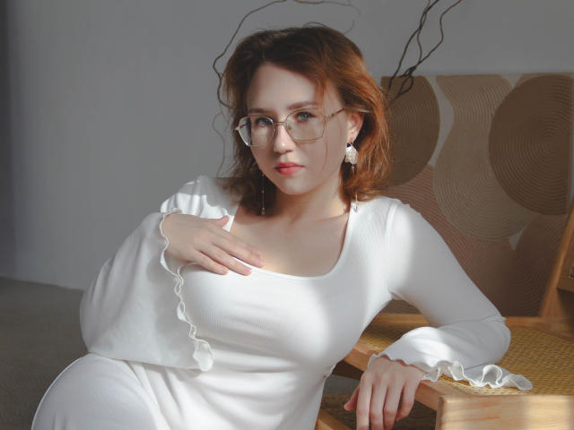 TeresaBirt with glasses, white dress, and earrings, has fair skin and red hair, resting hand on her tits, sitting in sunlit room, wooden chair with patterned cushion.