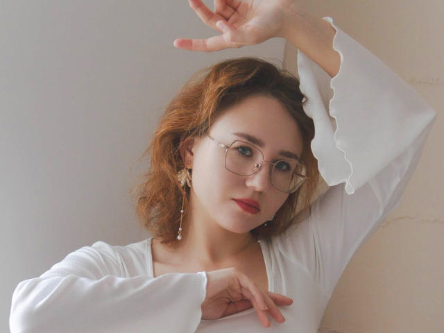 TeresaBirt with red hair, glasses, white long-sleeve top, and earrings, raising her right arm, looking at camera with red lips, soft light, pale skin, subtle makeup.
