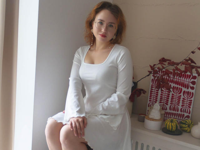 TeresaBirt with red hair, glasses, white dress, and small tits, sitting on windowsill, legs together, hands on knees, subtle smile, light skin, minimal makeup, soft light.