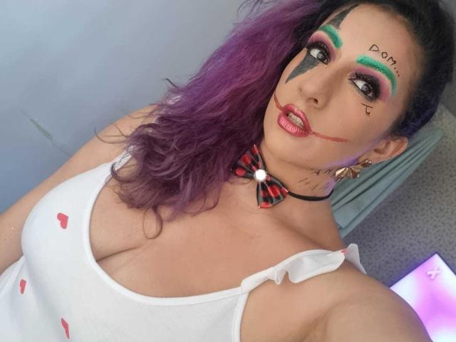 AndreaSharick with purple hair, white tank top showing big tits, green and pink makeup, black face paint, red lips, plaid bow, and a black choker.