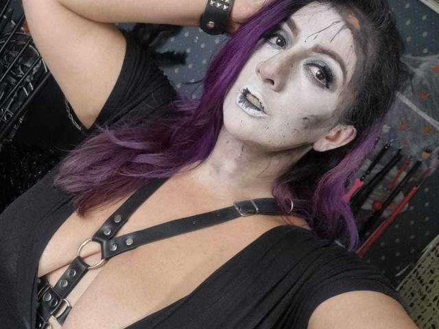 AndreaSharick with dark face paint, purple hair, black leather harness, and black top showing her tits. She's posing with one arm up, looking at the camera.