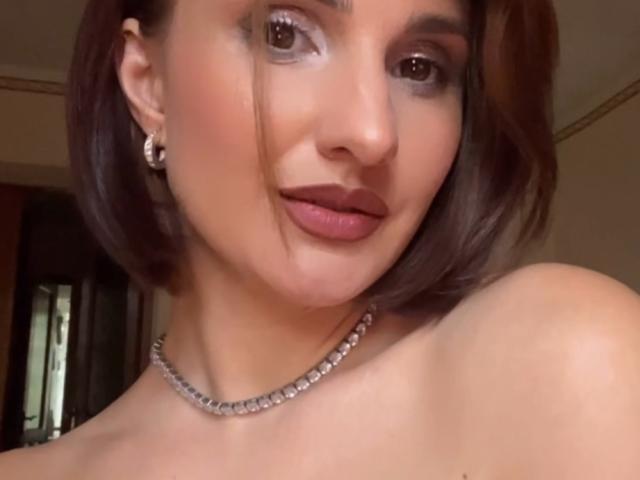 VeronicaLorie's face, short brown bob, wearing dark lipstick, diamond earrings, and a diamond necklace, smiling with a slight tilt to her head. Her tits are not visible.