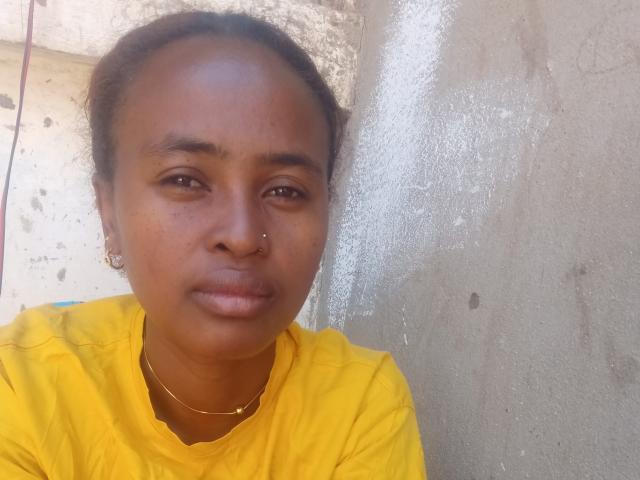 Lalance has medium brown skin, a nose ring, and a serious expression. She's wearing a yellow shirt and a gold necklace. Her hair is in a ponytail.