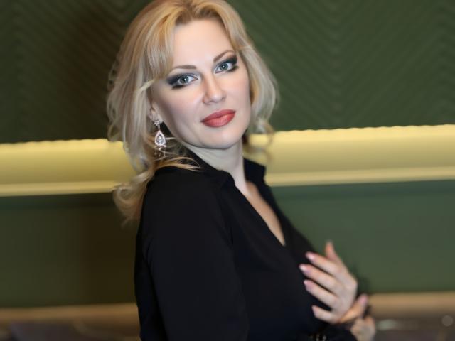 NatalyFoxy69 with blonde hair, blue eyes, and red lipstick, wearing a black top, hand on her tits, green and yellow background, looking at the camera with a slight smile.